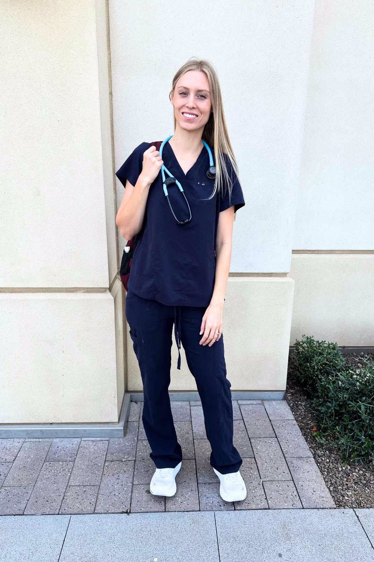A smiling nurse wearing a stethoscope, navy scrubs, and white shoes
